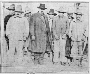 Group of many men in suits and hats. one white man (the marshal) in the middle of the Native American men.
