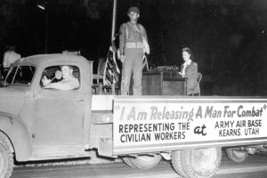 Woman typing and soldier standing on the back of a truck with the sign, "I Am Releasing a Man for Combat," Representing the civilian workers at Army Air Base Kearns Utah.